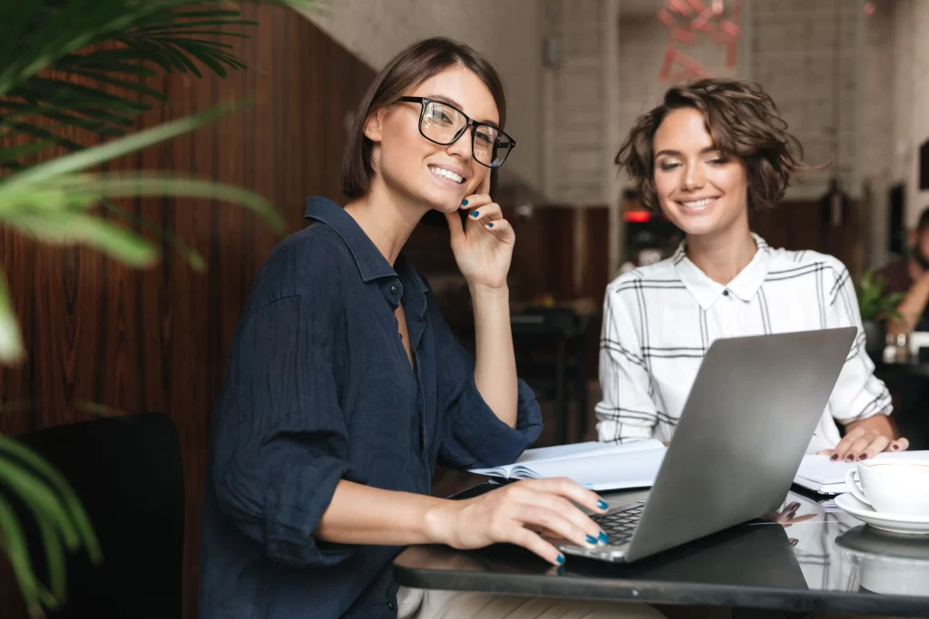 Duas mulheres sorrindo enquanto trabalham em um laptop em um ambiente moderno e acolhedor. Uma delas está usando óculos e parece engajada, sugerindo uma atmosfera de colaboração e produtividade.