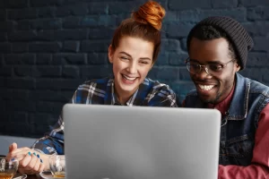 Dois colegas sorrindo enquanto colaboram em um projeto no computador. Estão em um ambiente descontraído e moderno, refletindo um momento de trabalho em equipe e aprendizado.