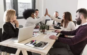 Seis colegas de trabalho em uma sala de reunião, duas delas fazendo um high five, representando cooperação e sucesso em equipe, enquanto os outros sorriem e observam.