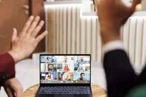 Duas pessoas acenando para uma tela de laptop que mostra uma videochamada com várias pessoas, simbolizando uma reunião virtual e trabalho remoto colaborativo.