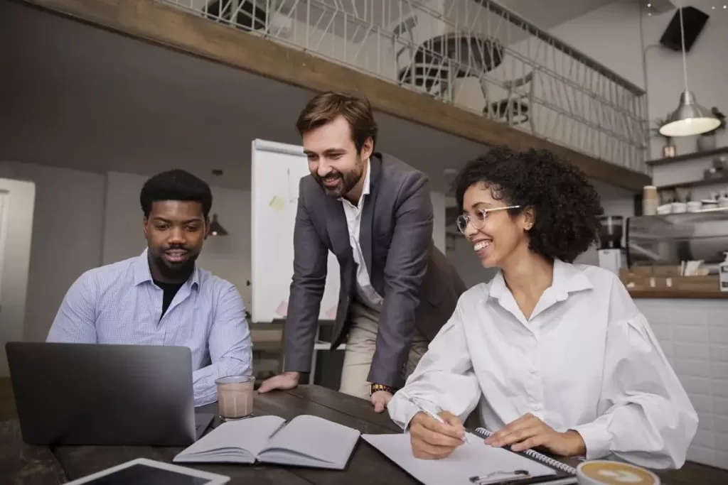 Três colegas de trabalho reunidos em uma mesa, sorrindo e colaborando em um projeto com um laptop e cadernos, em um ambiente de escritório moderno e descontraído.