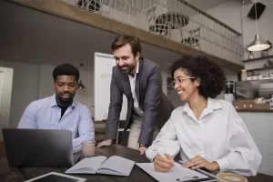 Três colegas de trabalho reunidos em uma mesa, sorrindo e colaborando em um projeto com um laptop e cadernos, em um ambiente de escritório moderno e descontraído.