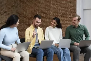 Quatro pessoas sentadas em cadeiras com laptops no colo, conversando e sorrindo em um ambiente descontraído com uma parede de tijolos ao fundo.