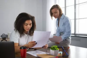 Duas mulheres analisando documentos em uma mesa de escritório, colaborando em um projeto, com materiais de trabalho e um copo de café ao redor.