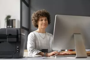 Mulher trabalhando em um computador de mesa em um escritório, concentrada e com expressão calma, com uma impressora ao lado.
