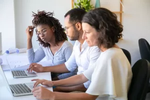 Três colegas de trabalho concentrados em laptops, colaborando lado a lado em uma mesa, em um ambiente de escritório iluminado e organizado.