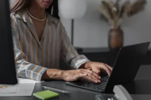 Mulher digitando em um laptop em sua mesa de trabalho, com materiais de escritório ao redor, em um ambiente elegante e bem decorado.