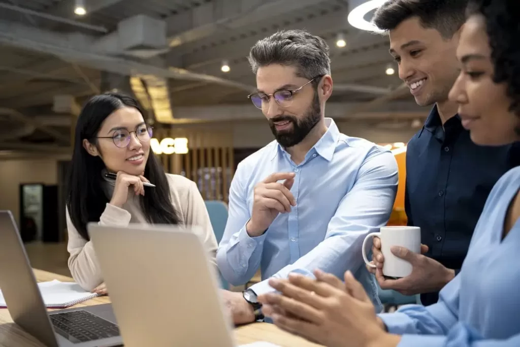 Quatro colegas de trabalho reunidos em frente a um laptop, discutindo e colaborando em um projeto, com sorrisos e interação em um ambiente de escritório moderno.