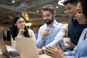 Quatro colegas de trabalho reunidos em frente a um laptop, discutindo e colaborando em um projeto, com sorrisos e interação em um ambiente de escritório moderno.
