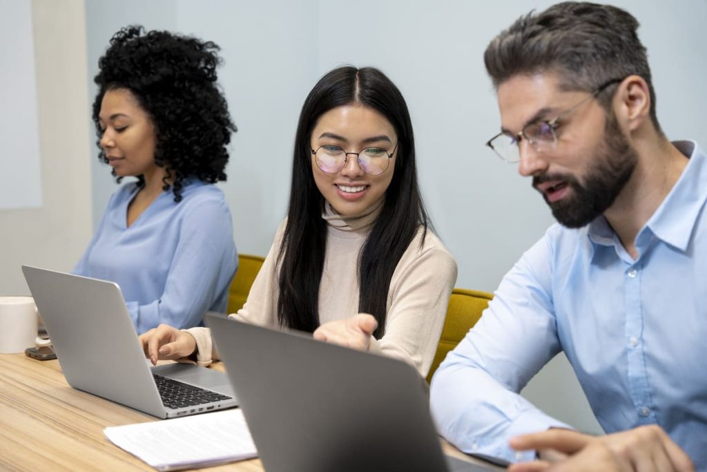 A imagem retrata um ambiente de trabalho colaborativo, onde três profissionais estão focados em suas atividades em laptops. A mulher no centro, com óculos e um sorriso, parece interagir com os colegas à sua frente, enquanto eles estão concentrados em suas telas. O cenário transmite uma atmosfera de produtividade e trabalho em equipe, com um design moderno e acolhedor.