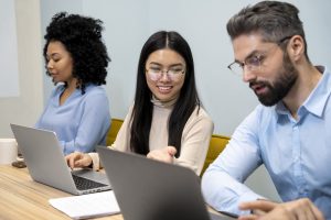 A imagem retrata um ambiente de trabalho colaborativo, onde três profissionais estão focados em suas atividades em laptops. A mulher no centro, com óculos e um sorriso, parece interagir com os colegas à sua frente, enquanto eles estão concentrados em suas telas. O cenário transmite uma atmosfera de produtividade e trabalho em equipe, com um design moderno e acolhedor.