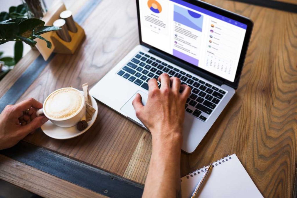 A imagem mostra uma cena de trabalho descontraída, onde uma pessoa está usando um laptop em uma mesa de madeira. A mão direita está no teclado, sugerindo que ela está digitando ou navegando, enquanto a mão esquerda segura uma xícara de café com espuma, transmitindo uma sensação de conforto e relaxamento. Há um caderno com uma caneta ao lado, indicando que a pessoa pode estar fazendo anotações ou planejando tarefas. A tela do laptop exibe uma interface que parece ser uma rede social ou plataforma de comunicação, reforçando a ideia de trabalho ou estudo em um ambiente casual e acolhedor. Além disso, a presença de itens como açúcar e um pequeno biscoito ao lado do café adiciona um toque acolhedor ao cenário.