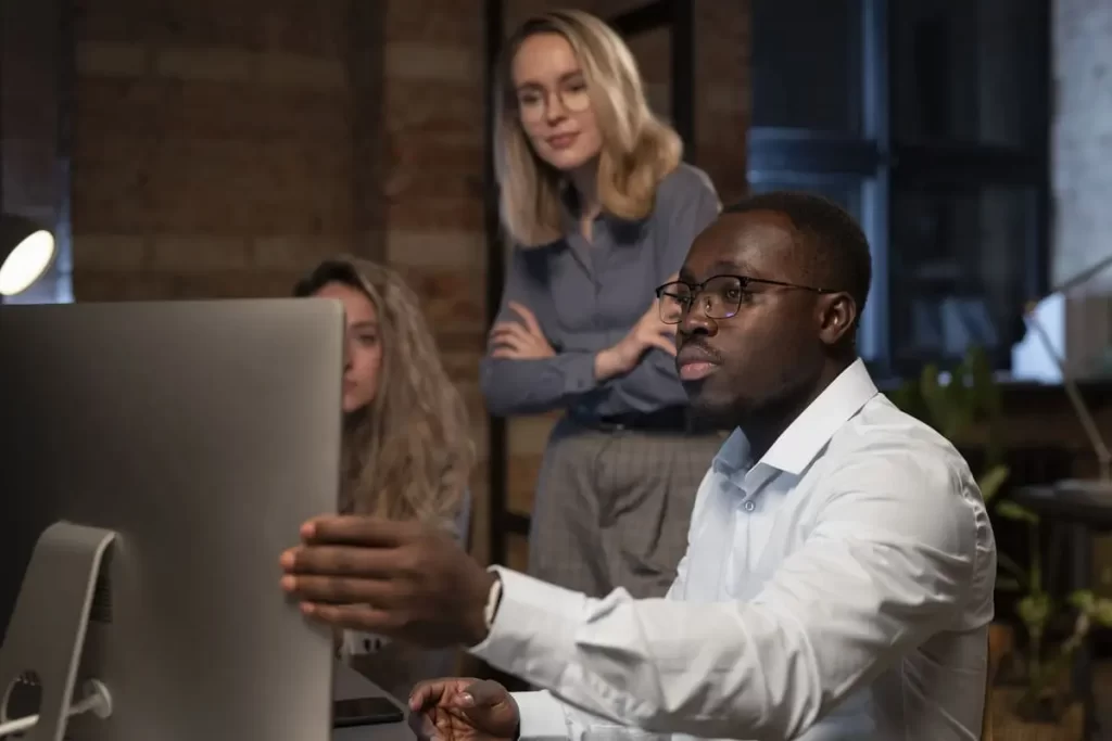 Três colegas de trabalho reunidos em frente a um computador, colaborando em um projeto durante o expediente, em um ambiente de escritório moderno e focado.