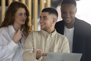 A imagem mostra três pessoas conversando de forma descontraída, aparentemente em um ambiente de trabalho colaborativo. Todos estão sorrindo e parecem engajados na conversa, sugerindo um clima amigável e de troca de ideias. O jovem no centro está gesticulando, o que indica que ele está explicando algo para os outros dois, que estão atentos. A cena reflete bem um ambiente de trabalho onde a comunicação e a colaboração são valorizadas, destacando a importância de uma equipe integrada e que se apoia mutuamente.