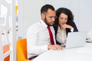 Um homem e uma mulher estão sentados em uma mesa de reunião, ambos concentrados em um laptop. O homem usa uma camisa branca e uma gravata vermelha, enquanto a mulher veste um blazer escuro sobre uma camisa clara. Eles parecem estar discutindo ou analisando algo no computador, demonstrando colaboração em um ambiente de trabalho moderno e bem iluminado.