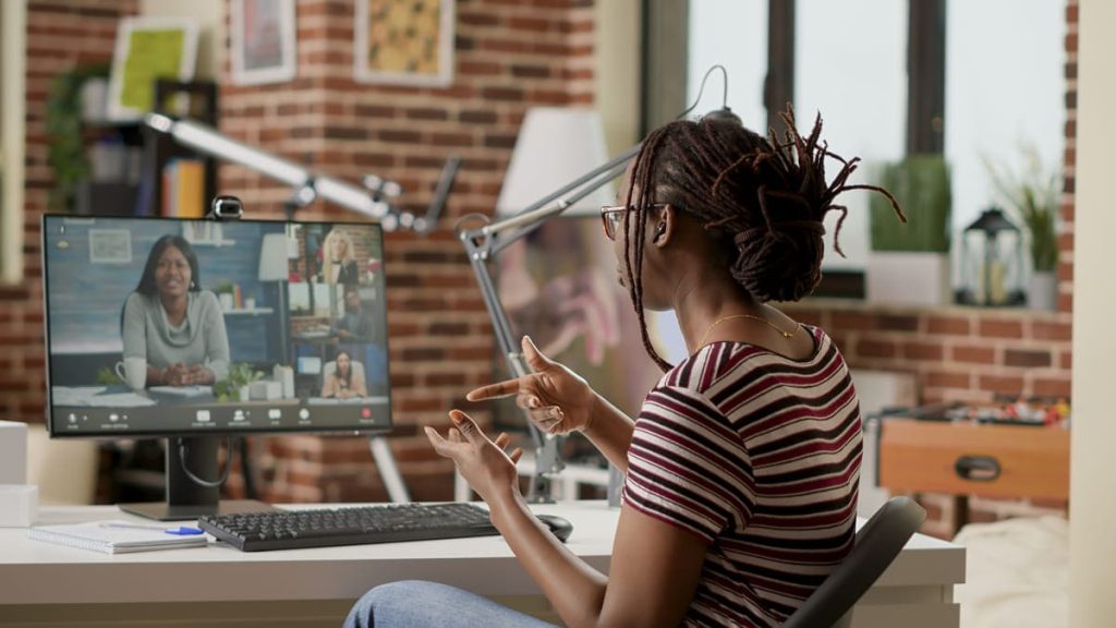 A imagem mostra uma mulher de costas participando de uma reunião virtual. Ela tem cabelo preso em tranças e está vestindo uma camiseta listrada em tons de vermelho e branco. A mulher está interagindo ativamente, gesticulando com as mãos enquanto observa um monitor na sua frente que exibe outras pessoas em uma videoconferência. Ao fundo, há uma parede de tijolos expostos e decoração de escritório, incluindo plantas e um objeto de jogo. O ambiente parece ser moderno e bem iluminado, indicando um espaço de trabalho colaborativo.