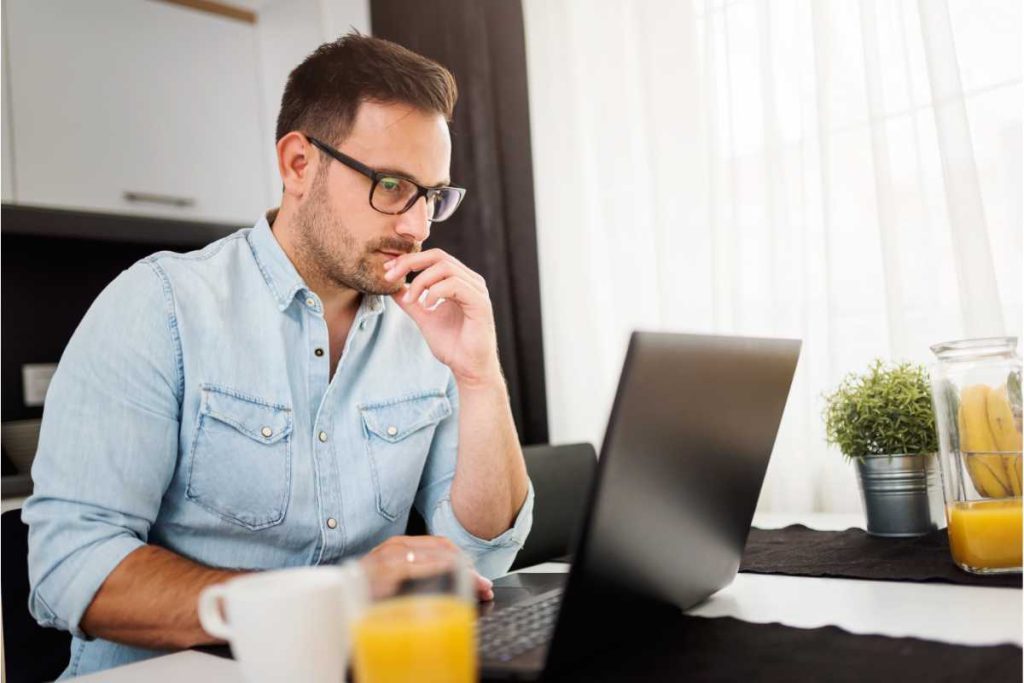 A imagem mostra um homem concentrado em frente ao laptop, sugerindo um momento de foco e trabalho. Ele está sentado em um ambiente iluminado, possivelmente uma cozinha ou sala de estar, com uma cortina branca que permite a entrada de luz natural suave. O homem usa óculos e está vestido de forma casual, com uma camisa jeans, o que transmite um tom descontraído, mas produtivo. Na mesa, há uma xícara de café e um copo de suco de laranja, além de um vaso com uma pequena planta e um pote com bananas, que contribuem para uma sensação de ambiente saudável e organizado. O cenário sugere uma rotina de home office confortável e equilibrada.