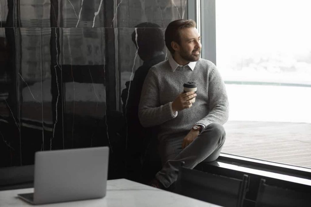 A imagem mostra um homem sentado em uma borda de janela, segurando um copo de café e olhando para fora. Ele está vestido de forma casual, com um suéter cinza e calça social, e aparenta estar relaxado ou pensativo, talvez aproveitando uma pausa. Ao fundo, vemos uma parede escura com um acabamento em mármore, o que dá um toque elegante e sofisticado ao ambiente. Em primeiro plano, há um laptop sobre a mesa, sugerindo que ele estava trabalhando antes de fazer uma pausa. A iluminação natural que entra pela janela cria uma atmosfera tranquila e contemplativa.