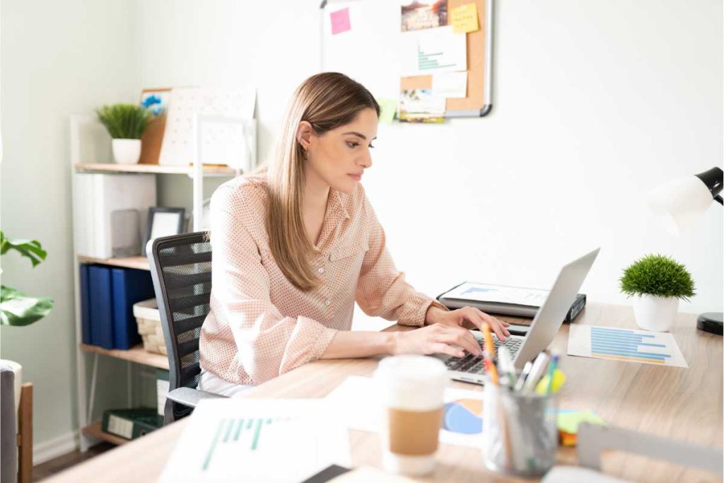 A imagem mostra uma mulher concentrada, trabalhando em seu laptop em um ambiente de escritório bem iluminado e organizado. Sobre a mesa, estão diversos itens como gráficos, um copo de café, canetas e uma pequena planta, sugerindo um ambiente de trabalho produtivo e acolhedor. Ao fundo, é possível ver uma estante com materiais e uma lousa de cortiça com notas e lembretes, indicando uma pessoa que valoriza a organização e planejamento. A imagem transmite uma atmosfera de foco e profissionalismo, destacando um momento típico de trabalho em um escritório moderno e bem arranjado.