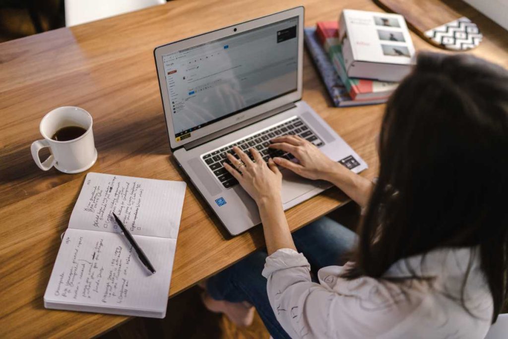 A imagem mostra uma pessoa trabalhando em um laptop, com foco em suas mãos digitando. Sobre a mesa de madeira, há um caderno aberto com anotações e uma caneta, sugerindo que a pessoa está organizando ideias ou realizando tarefas. Ao lado do caderno, há uma xícara de café, contribuindo para um ambiente de trabalho confortável e produtivo. Também são visíveis livros e outros objetos no fundo, criando uma atmosfera de estudo ou trabalho concentrado. A cena passa uma sensação de produtividade e organização, destacando um momento de foco e planejamento.