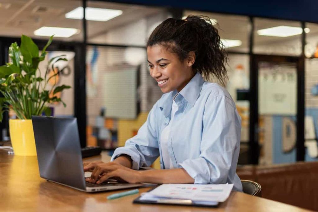 A imagem mostra uma mulher trabalhando em um laptop, sorrindo enquanto digita. Ela está vestindo uma camisa social azul clara e parece estar em um ambiente de trabalho moderno e descontraído, possivelmente um escritório ou coworking. Ao lado dela, há uma planta em um vaso amarelo, o que adiciona um toque de cor e natureza ao espaço. Também é possível ver papéis e uma caneta sobre a mesa, sugerindo que ela está organizando suas tarefas ou trabalhando em algum projeto. O ambiente ao fundo está desfocado, mas parece animado e bem iluminado, contribuindo para uma atmosfera acolhedora e produtiva.
