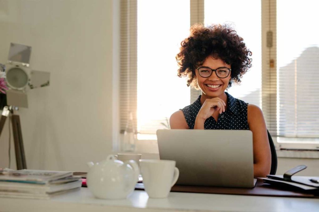 A imagem apresenta uma mulher sorridente, sentada em uma mesa de trabalho com um laptop à sua frente. Ela parece estar em um ambiente iluminado pela luz natural, com cortinas e persianas ao fundo que filtram a claridade, criando uma atmosfera agradável e confortável. Na mesa, há uma chaleira, xícaras de chá e algumas revistas ou papéis empilhados, sugerindo um ambiente descontraído e propício para produtividade. A expressão positiva e relaxada da mulher transmite confiança e alegria, refletindo um momento de satisfação no trabalho. O cenário combina elementos de um escritório moderno e acolhedor.