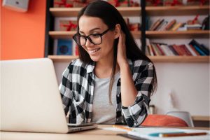 A imagem mostra uma mulher sorridente usando óculos e uma camisa xadrez preta e branca enquanto está sentada em uma mesa de trabalho. Ela parece concentrada em um laptop à sua frente, com uma mão atrás da cabeça, sugerindo um momento de descontração. Ao fundo, há uma estante com livros e alguns itens decorativos em uma parede de cor laranja. A atmosfera é acolhedora e moderna, ideal para um ambiente de trabalho criativo.