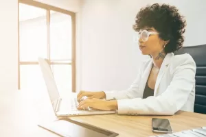 Retrato de uma mulher com cabelo encaracolado e óculos grandes, sentada em uma mesa de escritório, usando um blazer branco. Ela está digitando em um laptop, com um celular e outros materiais de escritório visíveis na mesa. A luz natural ilumina o ambiente, que possui janelas ao fundo.