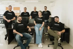 Time da Nubway - Um grupo de sete pessoas em um ambiente de escritório, todas usando camisetas pretas com o logotipo "nubiwag". As pessoas estão posando para a foto, com três em pé ao fundo e quatro sentadas na frente, criando uma atmosfera de equipe unida e colaborativa. O ambiente é iluminado e moderno, com computadores visíveis ao fundo.