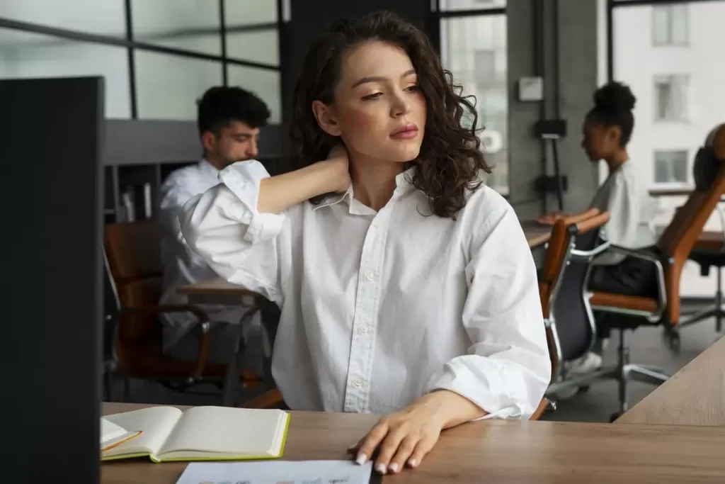 magem de uma mulher em um ambiente de escritório, com cabelo cacheado e vestido com uma camisa branca. Ela está sentada à mesa, segurando o pescoço com uma expressão que sugere cansaço ou desconforto. Ao fundo, outros colegas de trabalho estão visíveis, cada um em suas mesas. Um caderno aberto está à frente dela, indicando um ambiente de trabalho ativo.