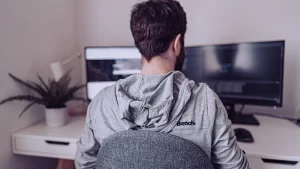 Um homem de costas, usando um moletom cinza, sentado em uma cadeira em frente a dois monitores. Ele está concentrado no trabalho, e há uma planta em um vaso ao lado da mesa.