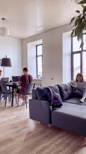 Três pessoas trabalhando em laptops, duas sentadas à mesa e uma relaxando no sofá em uma sala iluminada.