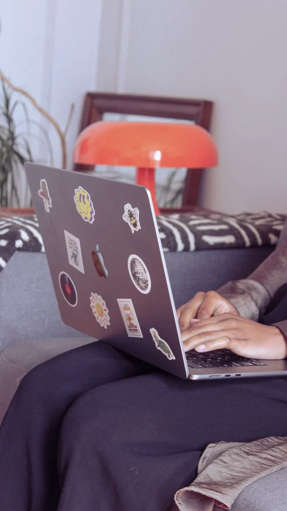 Pessoa trabalhando em um laptop com adesivos personalizados, em um ambiente aconchegante, sugerindo uma atmosfera criativa e informal.