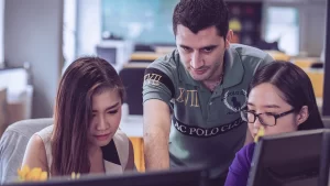 A imagem mostra três pessoas em um ambiente de trabalho, colaborando em frente a um computador. O homem no centro parece estar explicando algo para as duas mulheres ao lado, apontando para a tela. As expressões faciais sugerem concentração e atenção, indicando um momento de aprendizado ou troca de informações. O cenário ao fundo é de um escritório moderno, com luz suave e cadeiras confortáveis, criando um ambiente propício para trabalho em equipe e produtividade.