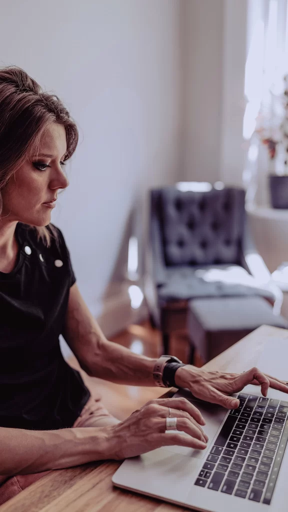 A imagem mostra uma mulher concentrada trabalhando em seu laptop em um ambiente iluminado e acolhedor. Ela está sentada em uma mesa de madeira, sugerindo um espaço de trabalho confortável e tranquilo. Ao fundo, há uma poltrona estofada e uma planta, que adicionam um toque de aconchego ao local. A iluminação natural que entra pela janela ajuda a criar uma atmosfera clara e inspiradora, ideal para o foco e a produtividade.