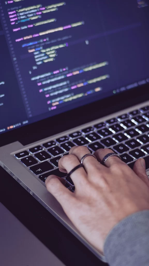 A imagem mostra uma mão digitando em um teclado de um laptop. A tela do laptop exibe código em um editor de texto, com sintaxe colorida, indicando que a pessoa está programando. A iluminação das teclas sugere um ambiente de pouca luz, criando um clima focado e concentrado. A mão na imagem usa anéis, adicionando um toque de estilo pessoal. A cena transmite a ideia de trabalho técnico e criação de software, associada a desenvolvimento de código e tarefas de programação.