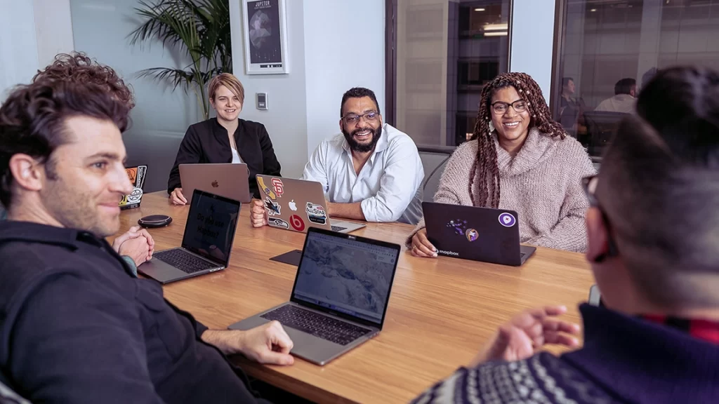 A imagem mostra um grupo de cinco pessoas reunidas em uma sala de reunião, cada uma com um laptop. Eles estão sentados ao redor de uma mesa de madeira e parecem engajados em uma discussão colaborativa, com sorrisos e expressões amigáveis. A cena transmite uma atmosfera descontraída e positiva, sugerindo um ambiente de trabalho inclusivo e aberto. A parede de vidro ao fundo reflete o espaço moderno do escritório, enquanto plantas e decoração minimalista contribuem para um cenário acolhedor. Os adesivos nos laptops adicionam um toque de personalidade, refletindo a diversidade de estilos e interesses dos participantes.