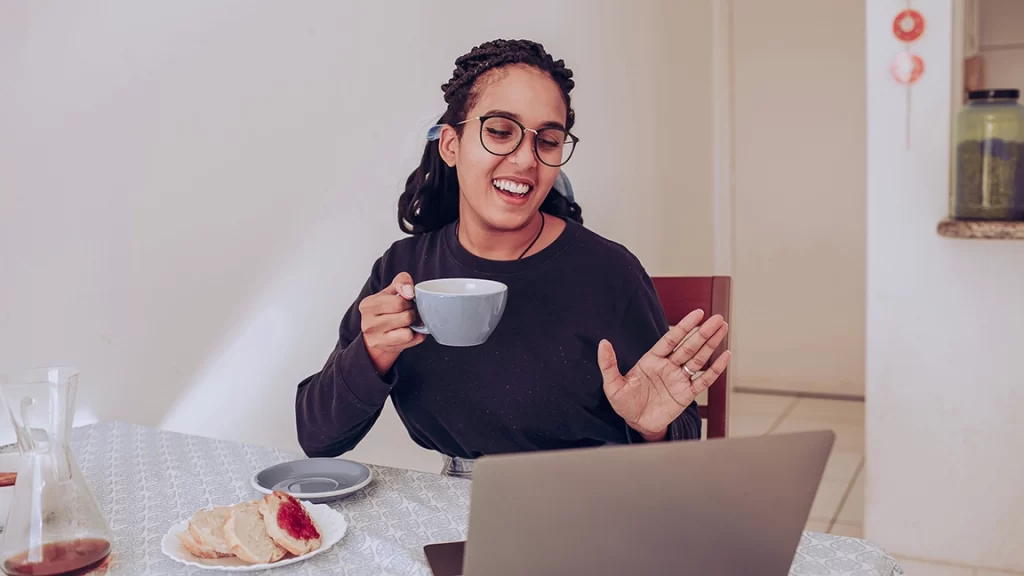 Uma mulher sorridente, segurando uma xícara de café, está participando de uma videochamada em um laptop, enquanto exibe um gesto de saudação. Ao fundo, há uma mesa com café, um prato com doces e uma decoração simples.