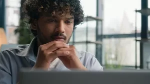 A imagem mostra um homem com cabelo encaracolado e um visual pensativo enquanto observa a tela do seu laptop. Ele está sentado em um ambiente moderno e bem iluminado, com grandes janelas ao fundo, sugerindo que ele pode estar refletindo sobre uma decisão ou focado em uma tarefa importante. O cenário transmite uma sensação de concentração e análise, possivelmente relacionado ao trabalho ou a uma situação que exige atenção e resolução.