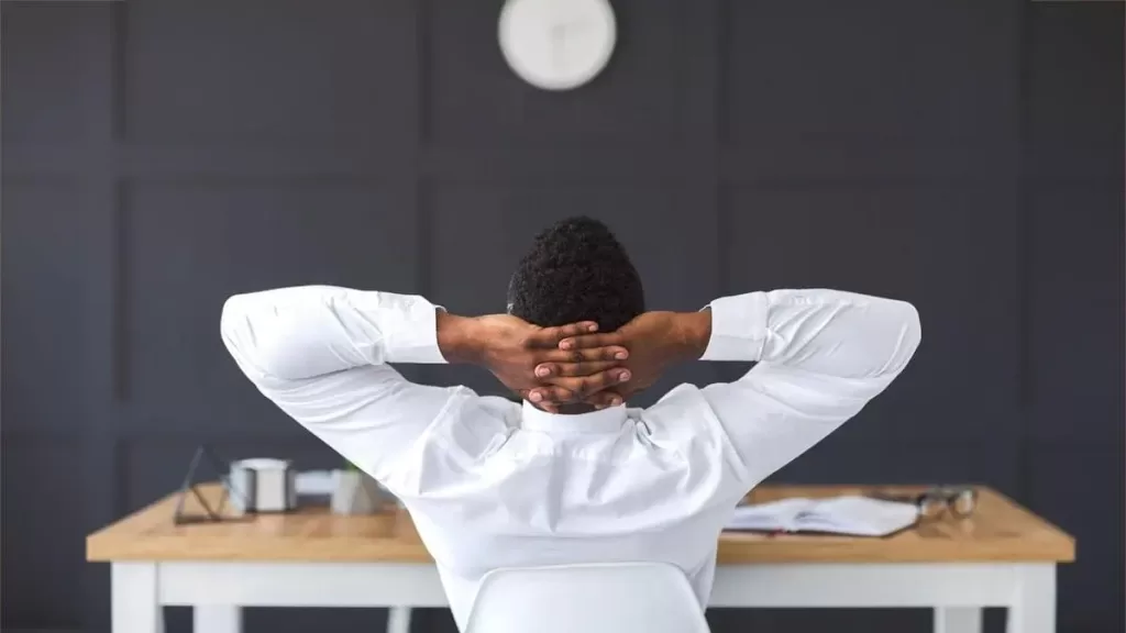 A imagem apresenta um homem sentado em uma cadeira de escritório, com as mãos entrelaçadas atrás da cabeça, descansando e olhando para a parede à sua frente. Ele está usando uma camisa social branca e parece estar em um ambiente de trabalho moderno, com um design minimalista e um relógio na parede. A cena transmite uma sensação de alívio ou relaxamento após um período de trabalho intenso, possivelmente refletindo sobre uma tarefa concluída ou fazendo uma pausa. O cenário é tranquilo e com uma iluminação suave, criando uma atmosfera relaxante.