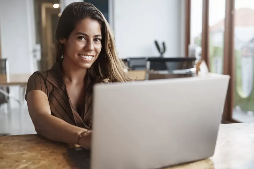 A imagem retrata uma mulher sorridente, sentada em uma mesa de madeira, utilizando um laptop. Ela tem cabelos longos e castanhos, e está vestindo uma blusa de manga curta com listras. O ambiente é bem iluminado, com janelas amplas ao fundo, sugerindo um espaço de trabalho moderno e aconchegante. A expressão dela indica satisfação e concentração, sugerindo que está envolvida em uma tarefa relacionada ao trabalho ou a estudos. Essa cena pode evocar temas de produtividade e a importância da tecnologia no cotidiano profissional.