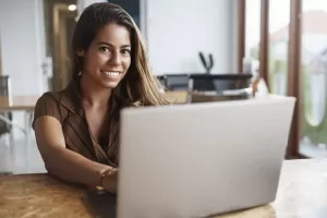 A imagem retrata uma mulher sorridente, sentada em uma mesa de madeira, utilizando um laptop. Ela tem cabelos longos e castanhos, e está vestindo uma blusa de manga curta com listras. O ambiente é bem iluminado, com janelas amplas ao fundo, sugerindo um espaço de trabalho moderno e aconchegante. A expressão dela indica satisfação e concentração, sugerindo que está envolvida em uma tarefa relacionada ao trabalho ou a estudos. Essa cena pode evocar temas de produtividade e a importância da tecnologia no cotidiano profissional.