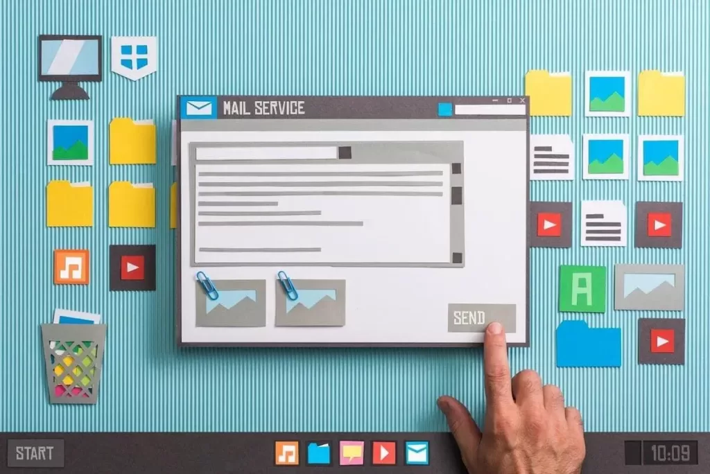 A imagem mostra uma composição artística com elementos em papel que simulam uma interface de serviço de e-mail. O foco está em uma tela de computador fictícia com uma caixa de e-mail composta por uma mensagem e opções de anexos, com ícones representando imagens. Também há diversos ícones de arquivos e aplicativos (como música, vídeo, imagem e documentos) ao redor, dando uma sensação de organização digital. A mão está prestes a clicar no botão "SEND" (enviar), simulando a ação de envio de e-mail. O design é feito com recortes de papel, dando um toque criativo e divertido à imagem