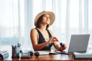 A imagem mostra uma mulher sentada em uma mesa, com um laptop à sua frente, olhando para cima enquanto segura um objeto com uma mão. Ela usa um chapéu de palha e está em um ambiente luminoso, com cortinas claras ao fundo. A cena sugere um momento de reflexão ou pausa enquanto ela trabalha, possivelmente em uma situação de férias ou de trabalho remoto. É um ambiente descontraído, ideal para tarefas mais criativas ou de planejament